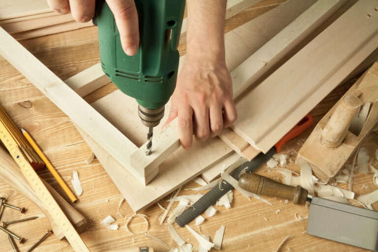 A person drilling into a piece of wood with an electric drill, surrounded by woodworking tools and wood scraps.